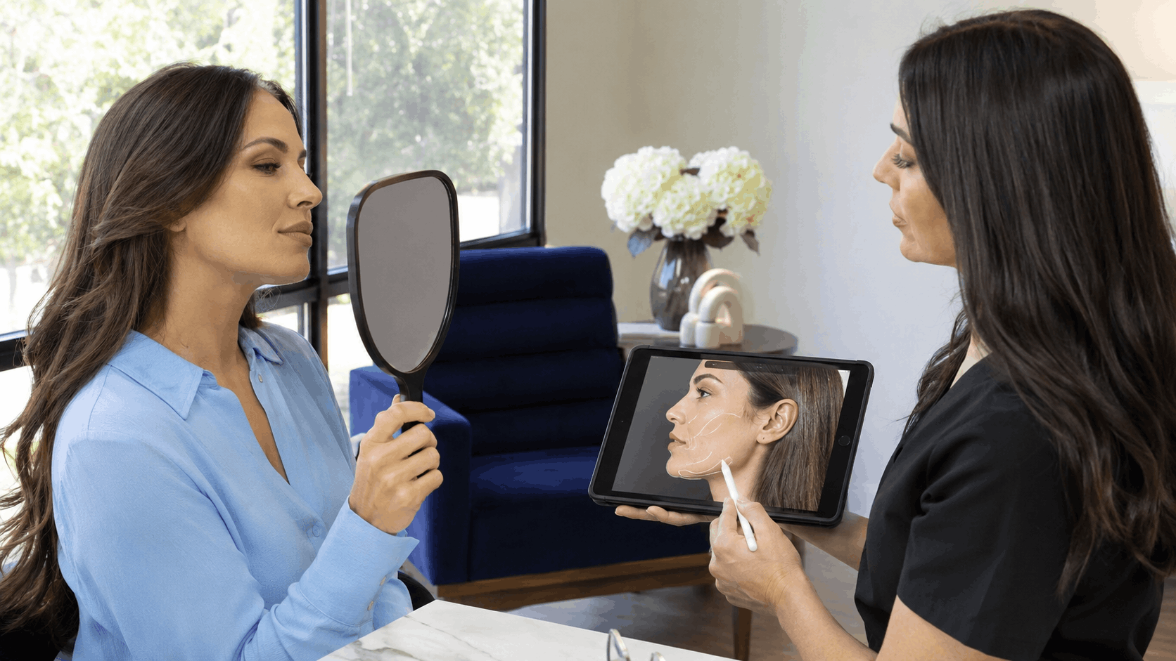 provider uses a tablet to illustrate facial contouring treatment areas while a patient looks in a hand mirror.
