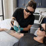provider using a laser device to treat a patient's tattooed forearm while both wear protective eyewear.
