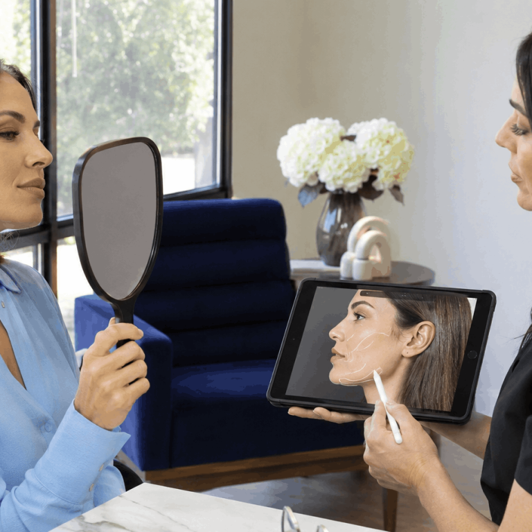 Provider uses a tablet to illustrate facial contouring treatment areas while a patient looks in a hand mirror.